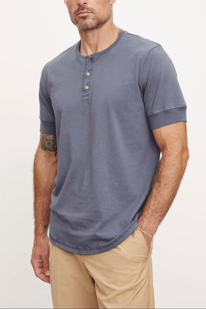 A man with a beard and tattoo on his arm, wearing a gray DEON HENLEY shirt with Henley details and beige pants, stands against a white background, looking at the camera.