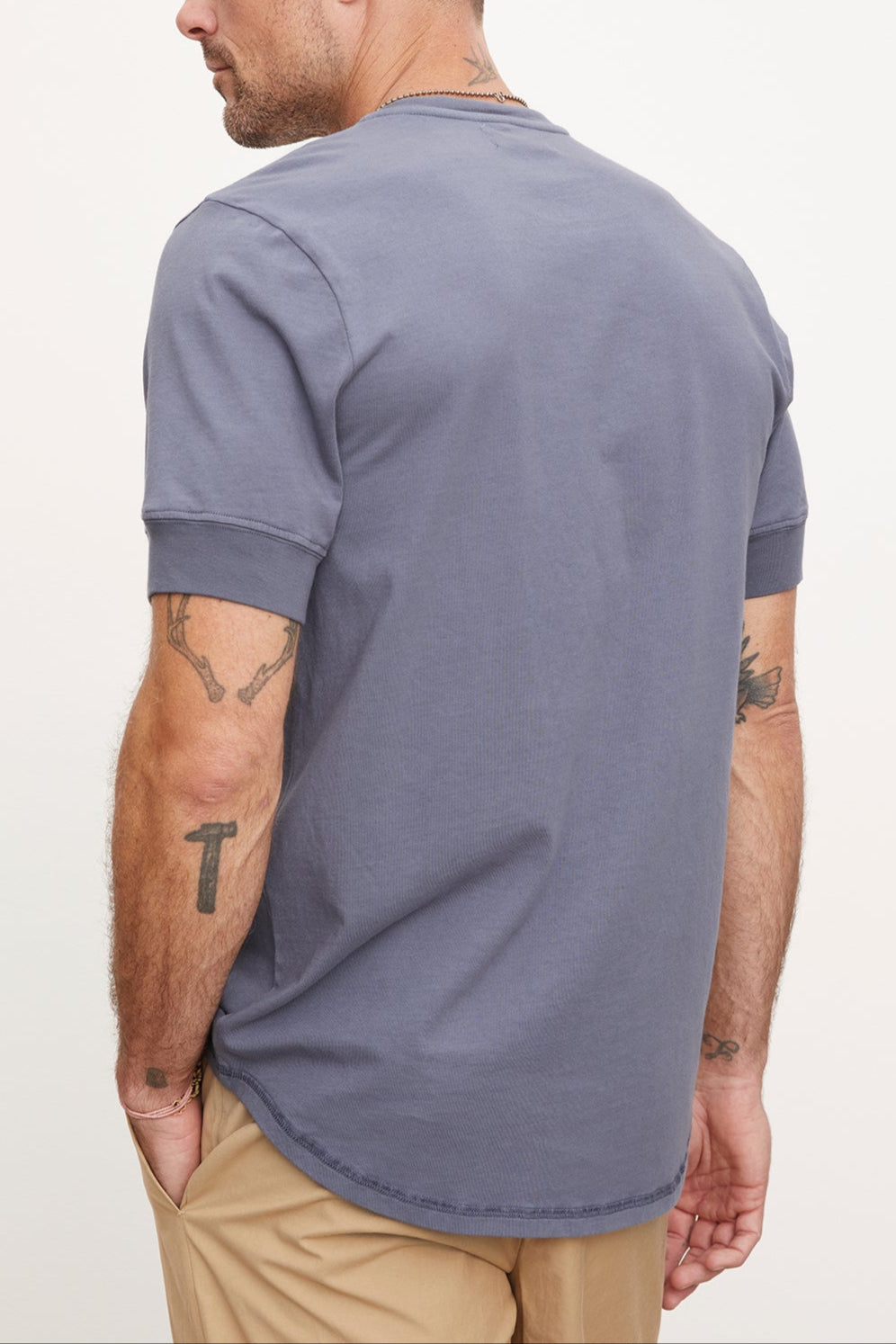 A man in a Velvet by Graham & Spencer DEON Henley t-shirt with scooped hem and beige pants, viewed from the side as he looks to the left, displaying tattoos on his arms.-36918612000961