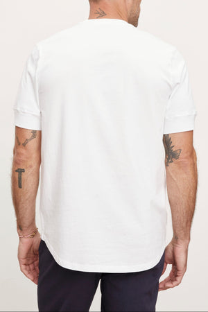 A man viewed from behind wearing a plain white short-sleeved Velvet by Graham & Spencer DEON HENLEY t-shirt and navy trousers, with visible tattoos on both arms.