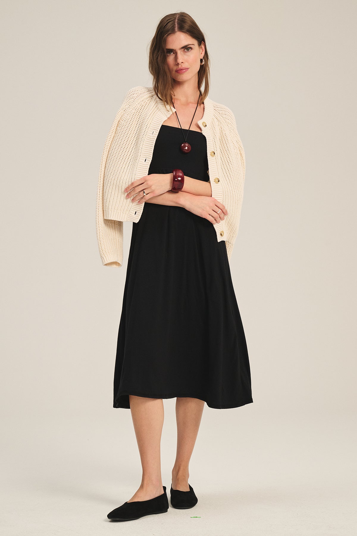 A woman stands against a plain background wearing the Velvet by Graham & Spencer LISABETH CARDIGAN draped over her shoulders, paired with a black midi dress, chunky necklace, and black flats.
