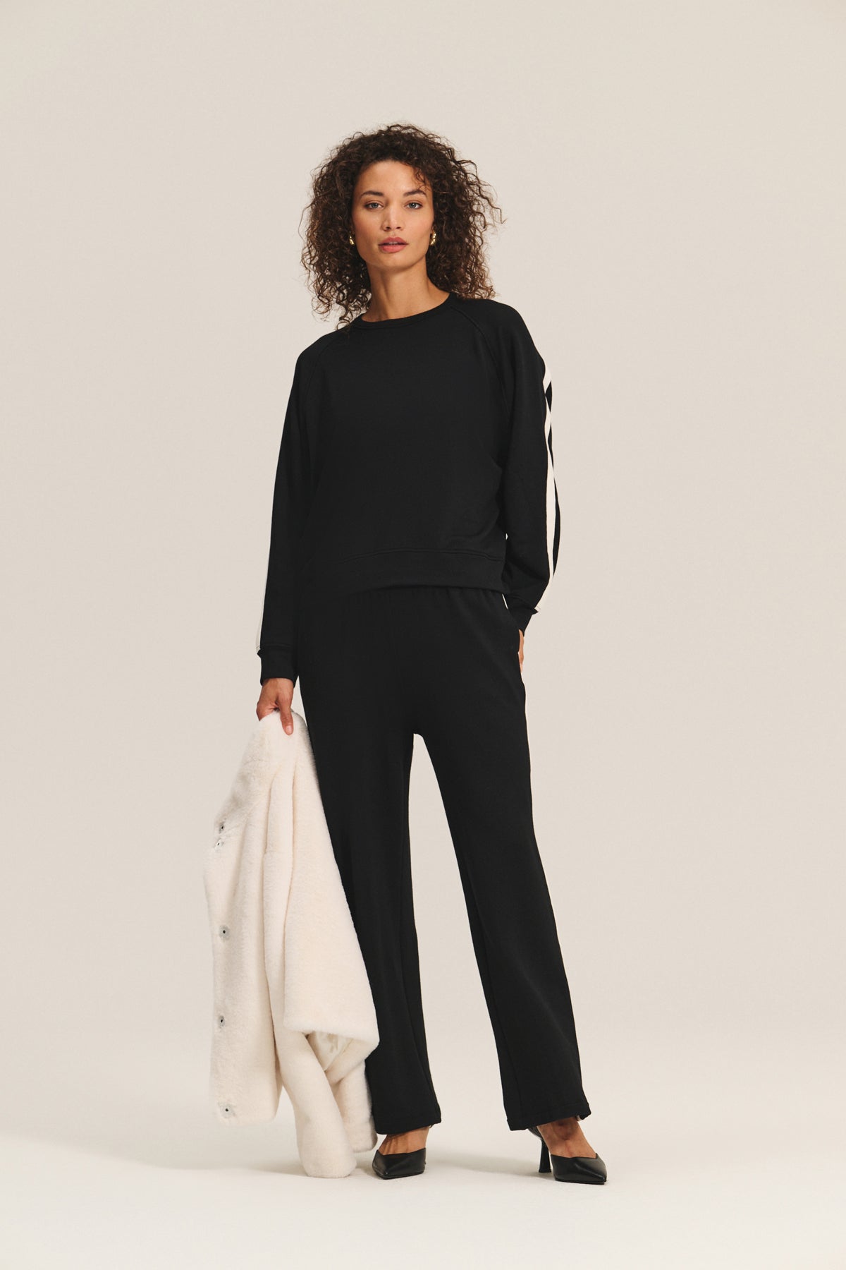 A woman holds the SHEENA LUXE FAUX FUR JACKET by Velvet by Graham & Spencer in her left hand, wearing a black long-sleeve top and matching pants, against a plain light background.-40234935025857