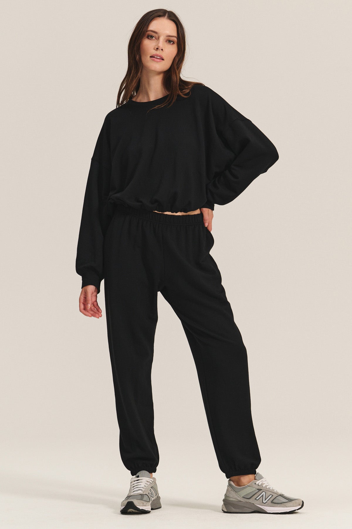 A woman wears the EVERS JOGGER by Velvet by Graham & Spencer—relaxed fit black fleece joggers and a matching sweatshirt—paired with gray New Balance sneakers, standing against a plain light background.-40334207156417