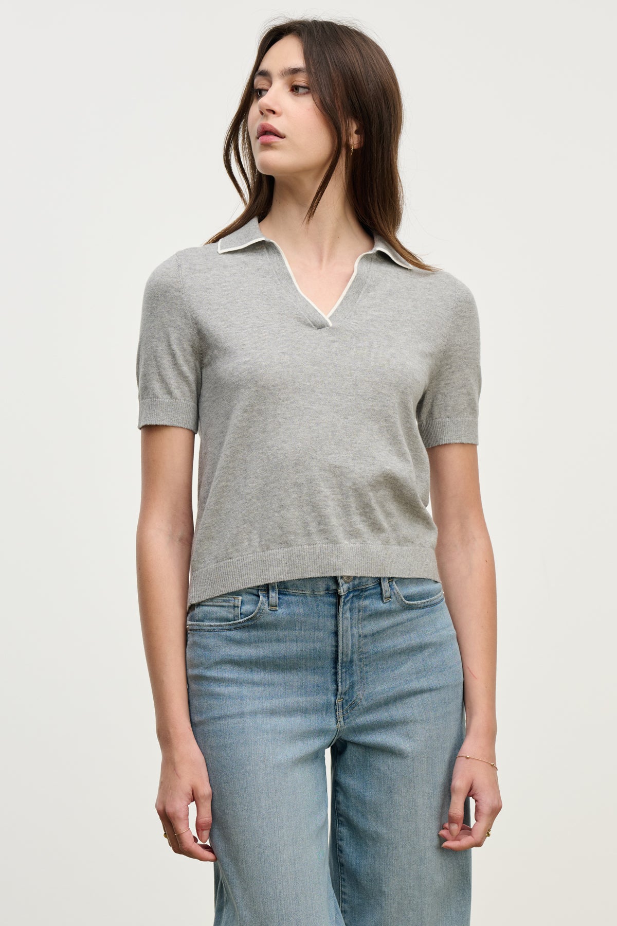 A woman stands against a plain light background wearing the ABELA SWEATER POLO by Velvet by Graham & Spencer, a light gray cropped sweater with a collar, paired with light blue jeans.-40046111785153