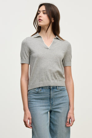 A woman stands against a plain light background wearing the ABELA SWEATER POLO by Velvet by Graham & Spencer, a light gray cropped sweater with a collar, paired with light blue jeans.