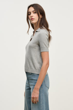A woman stands in profile against a plain background, wearing the ABELA SWEATER POLO by Velvet by Graham & Spencer—a modern gray cropped sweater polo with short sleeves—paired with classic blue jeans.