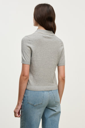 A woman faces away, wearing the ABELA SWEATER POLO by Velvet by Graham & Spencer with high-waisted blue jeans against a plain light background.