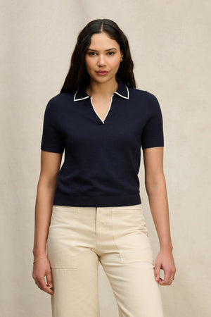 A woman stands against a neutral background, wearing the ABELA Cotton Cashmere Sweater Polo by Velvet by Graham & Spencer in navy with white trim, paired with beige pants.