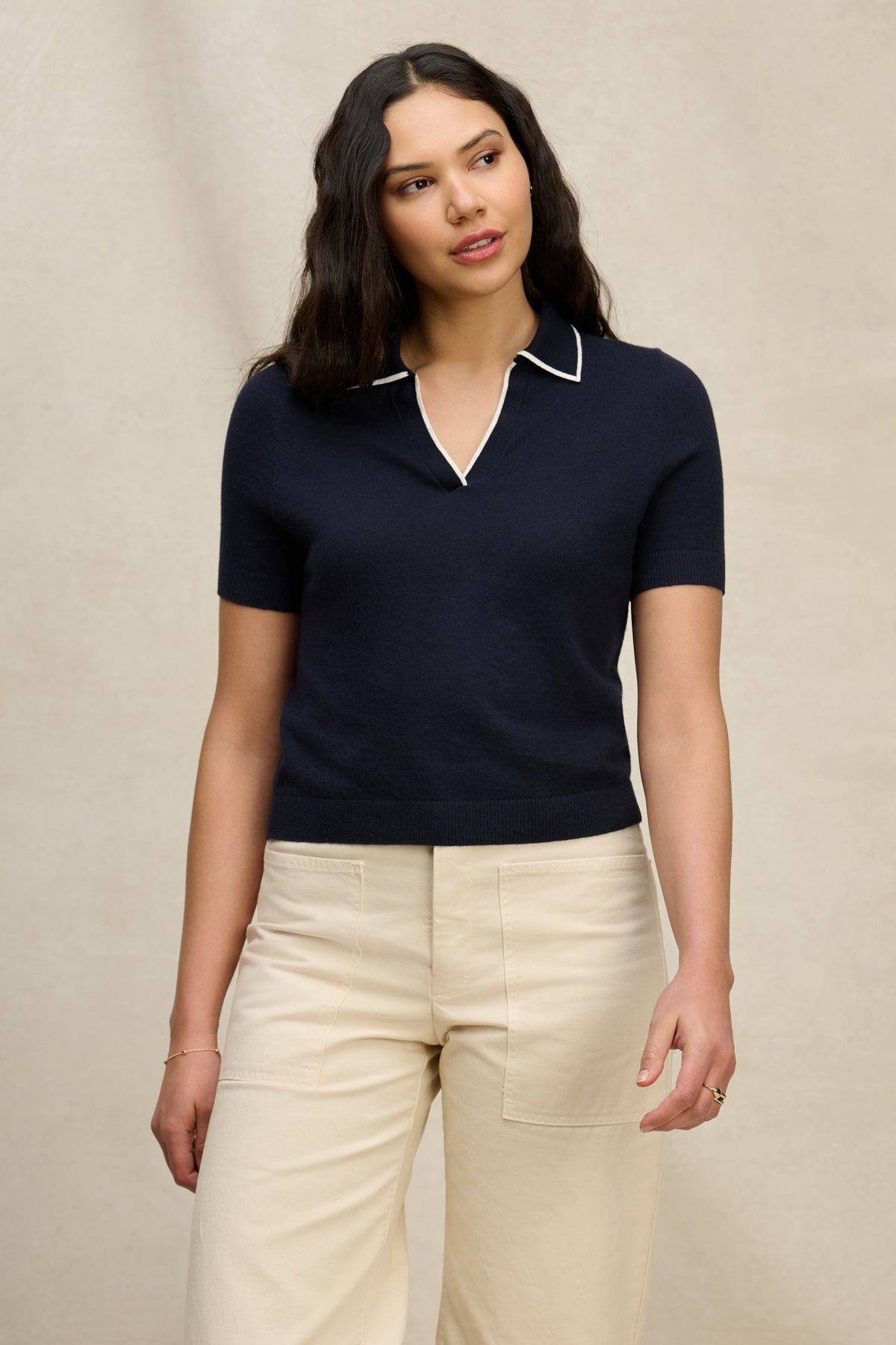 A woman with long dark hair wears the ABELA SWEATER POLO by Velvet by Graham & Spencer, a navy cropped polo with white trim, paired with cream-colored pants against a plain beige background.-39725305954497