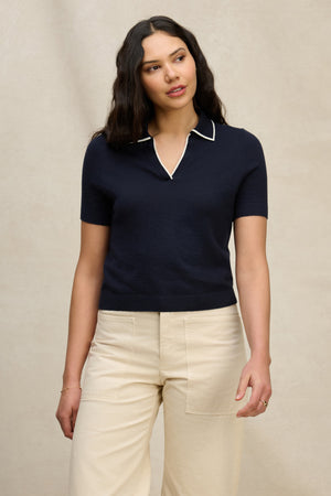 A woman with long dark hair wears the ABELA COTTON CASHMERE SWEATER POLO by Velvet by Graham & Spencer and high-waisted beige pants, standing against a plain light background.
