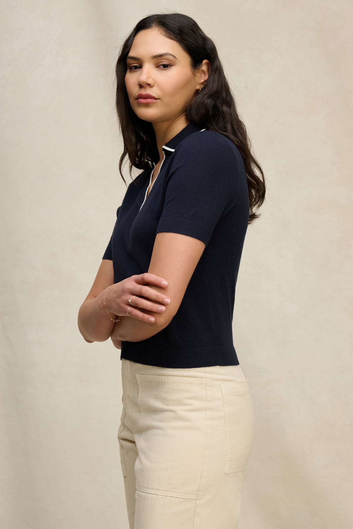 A woman with long brown hair stands with arms crossed, wearing the ABELA SWEATER POLO by Velvet by Graham & Spencer in navy and beige pants against a plain, light background.-40046111686849