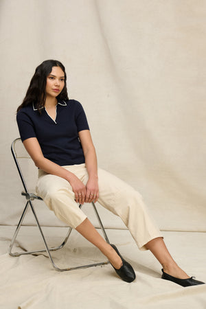 A woman sits on a metal chair against a beige backdrop, wearing the ABELA COTTON CASHMERE SWEATER POLO by Velvet by Graham & Spencer, paired with light beige pants and black shoes.