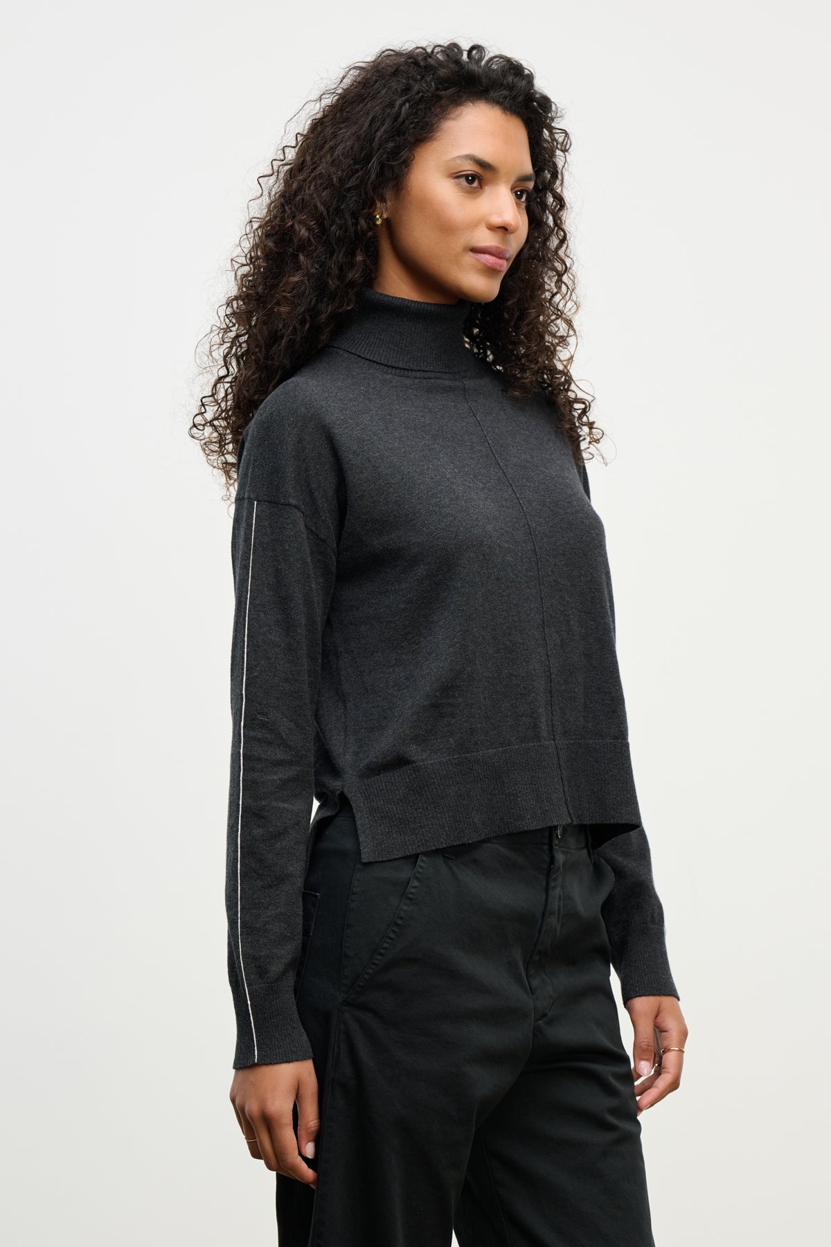 A woman with curly hair wears the LIBRA SWEATER by Velvet by Graham & Spencer— a dark gray cotton cashmere turtleneck—paired with dark pants, standing against a plain light background.-40046144094401