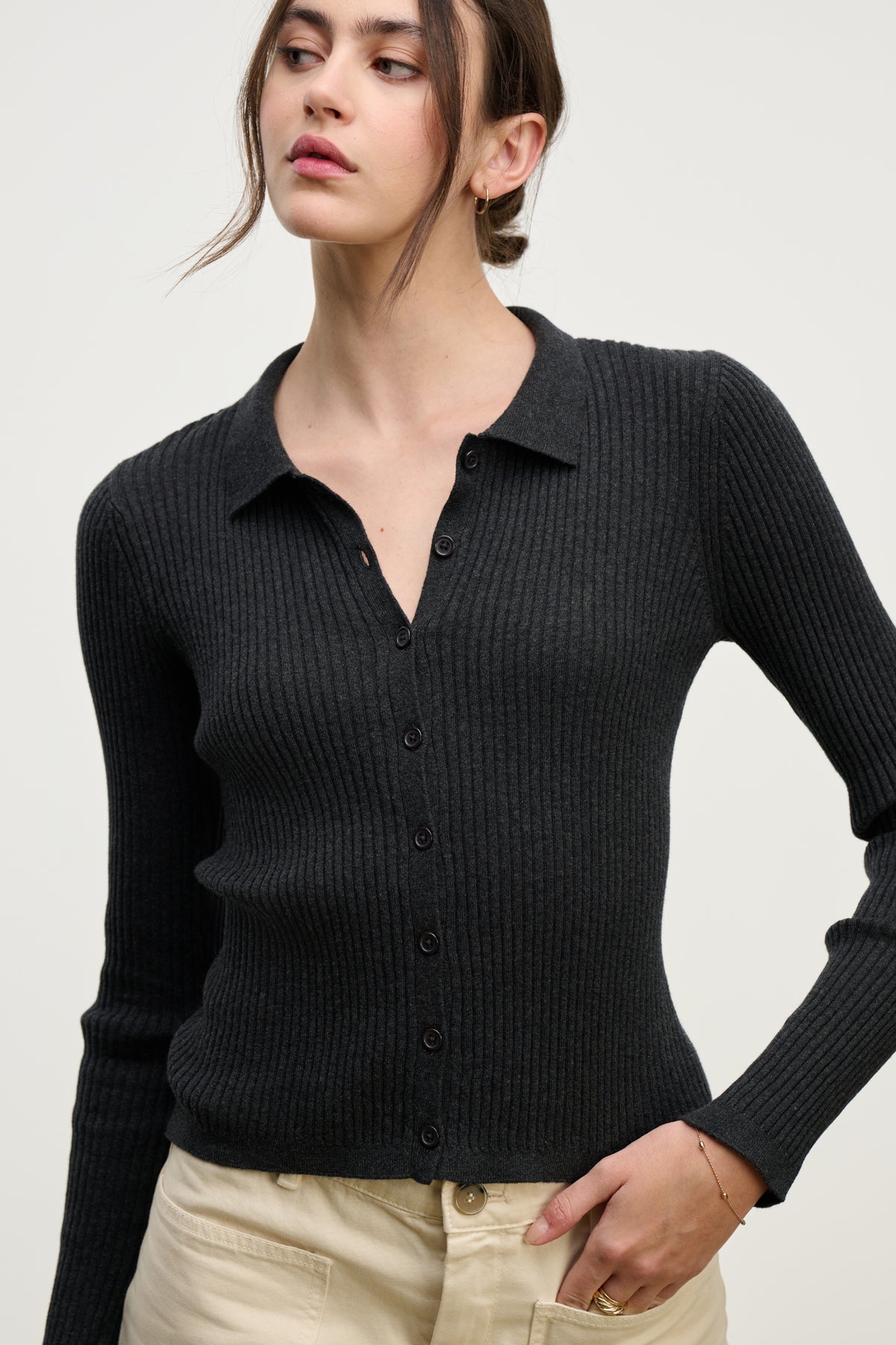 A woman wears the NIKKI COTTON CASHMERE CARDIGAN by Velvet by Graham & Spencer, a fitted ribbed black button-up with long sleeves, paired with light beige pants against a plain light background.