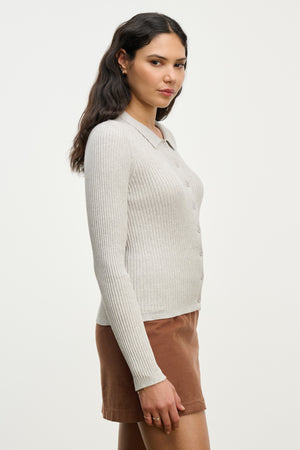 A woman in profile wears the NIKKI COTTON CASHMERE CARDIGAN by Velvet by Graham & Spencer, a light gray fitted layering piece, paired with a brown skirt against a plain white background.