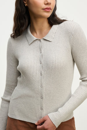 A woman wears the NIKKI COTTON CASHMERE CARDIGAN by Velvet by Graham & Spencer—a light gray, ribbed, fitted cardigan with a collar and long sleeves—paired with brown pants for versatile layering.
