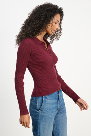 A woman stands in profile against a white background, wearing the Velvet by Graham & Spencer NIKKI COTTON CASHMERE CARDIGAN in maroon with blue jeans.