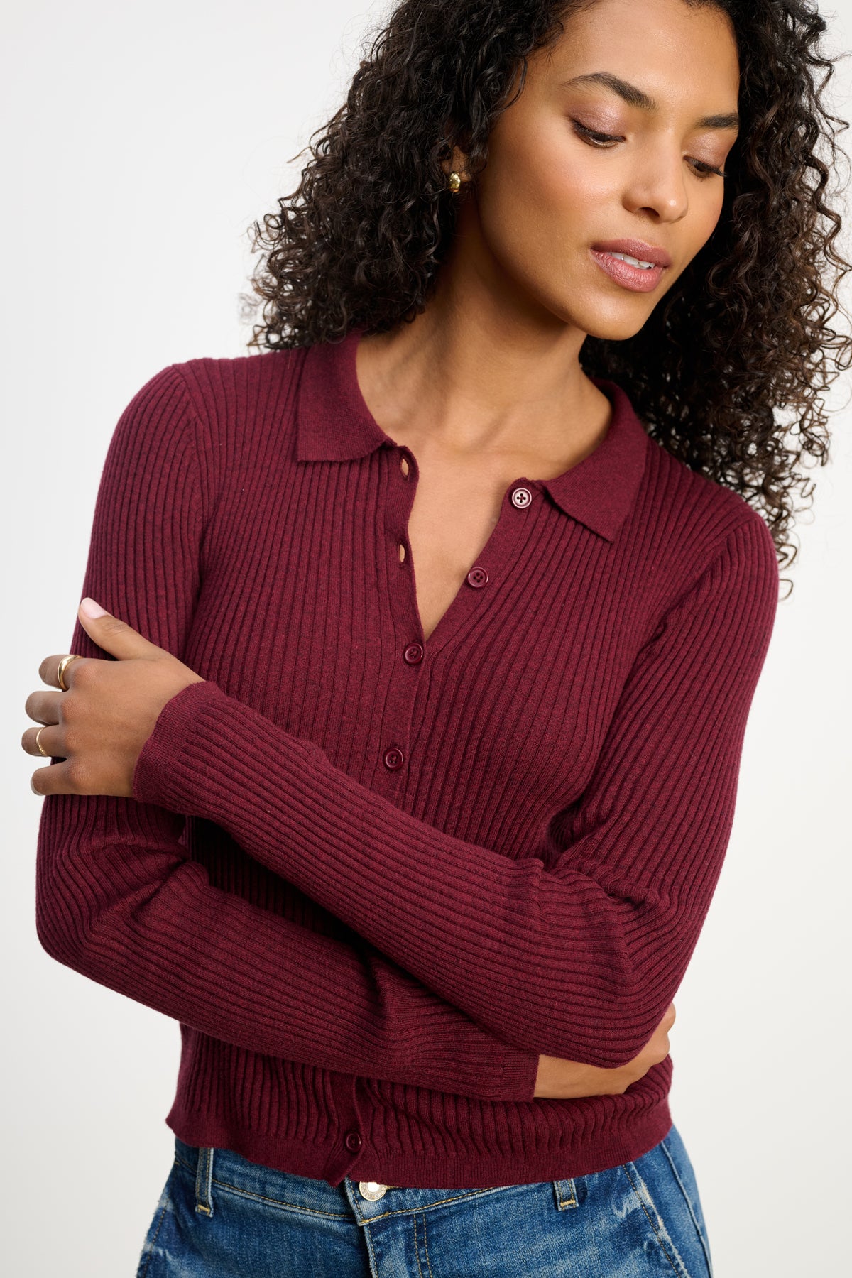 A woman with curly hair wears the Velvet by Graham & Spencer NIKKI COTTON CASHMERE CARDIGAN in burgundy, paired with blue jeans, standing with arms crossed and looking down.