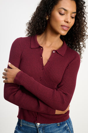 A woman with curly hair wears the Velvet by Graham & Spencer NIKKI COTTON CASHMERE CARDIGAN in burgundy, paired with blue jeans, standing with arms crossed and looking down.