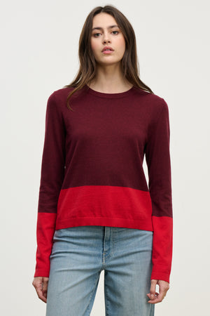 A woman wears a Velvet by Graham & Spencer ROXY SWEATER in burgundy and red with light blue jeans, standing against a plain light background. This lightweight sweater brings effortless style to any casual outfit.