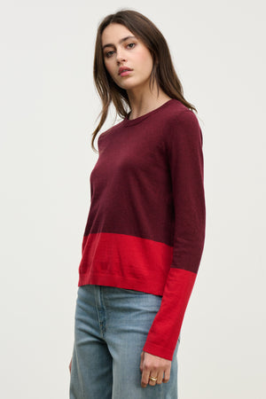 A woman wearing the ROXY SWEATER by Velvet by Graham & Spencer—a long-sleeve, burgundy and red color-block lightweight sweater—with blue jeans stands against a plain light background.