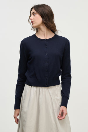 A woman wearing the VIOLETTE CARDIGAN by Velvet by Graham & Spencer—a navy blue, long-sleeve, button-up cotton cashmere cardigan—paired with a light skirt stands against a plain background, looking to her left.