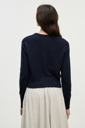 A person with long brown hair is shown from the back against a plain light background, wearing the VIOLETTE CARDIGAN by Velvet by Graham & Spencer in navy blue cotton cashmere and an off-white skirt.