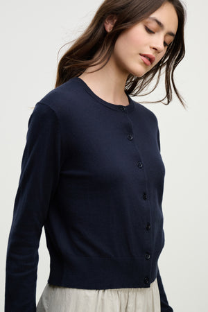 A person wearing the VIOLETTE CARDIGAN by Velvet by Graham & Spencer in navy blue with a light-colored skirt stands against a plain, light background.
