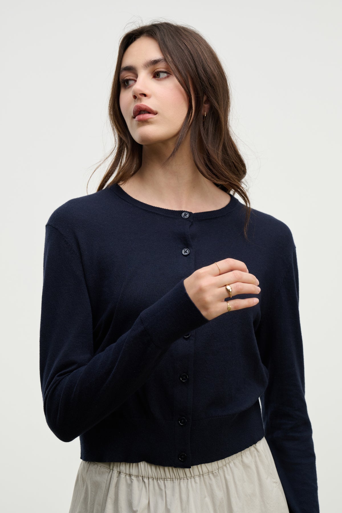 A woman with long brown hair wears the VIOLETTE CARDIGAN by Velvet by Graham & Spencer in navy blue, paired with a light beige skirt, standing against a plain light background.