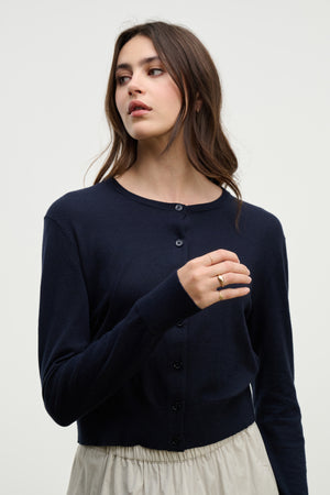 A woman with long brown hair wears the VIOLETTE CARDIGAN by Velvet by Graham & Spencer in navy blue, paired with a light beige skirt, standing against a plain light background.