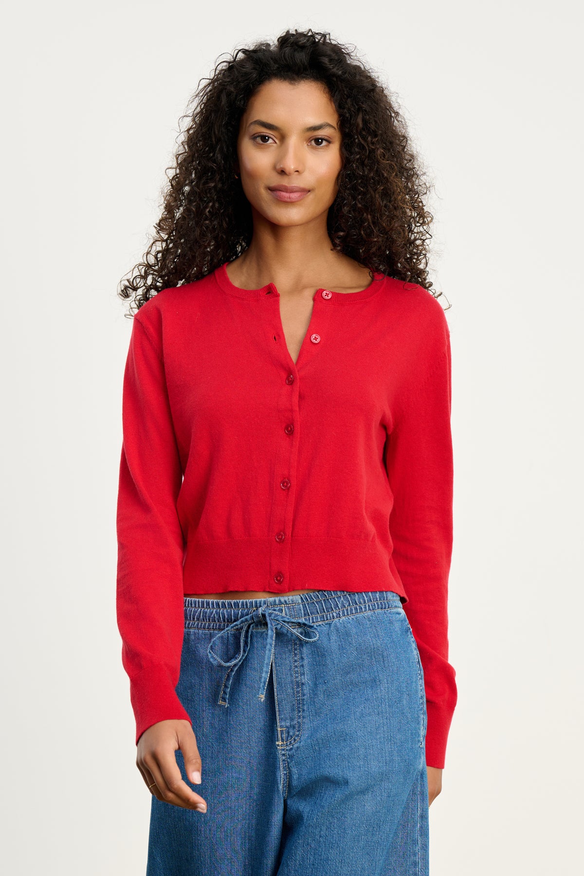A woman with curly hair wears the VIOLETTE CARDIGAN by Velvet by Graham & Spencer and blue drawstring jeans, standing against a plain light background.