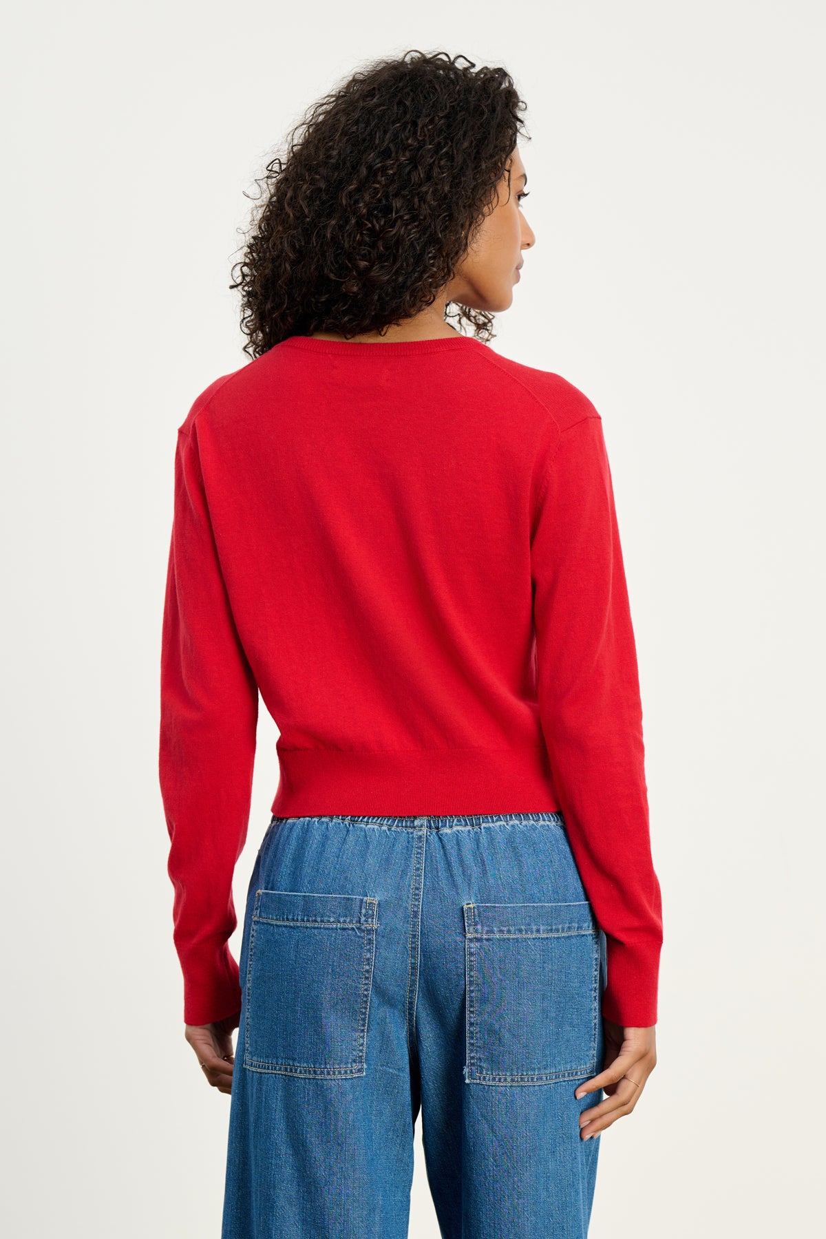A person with curly hair, wearing the VIOLETTE CARDIGAN by Velvet by Graham & Spencer in red and blue jeans, stands with their back to the camera against a plain background.