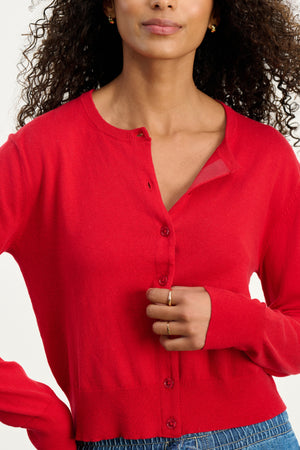 A woman with curly hair, gold earrings, and a ring on her left hand wears the VIOLETTE CARDIGAN by Velvet by Graham & Spencer in red with blue jeans, standing against a plain background.