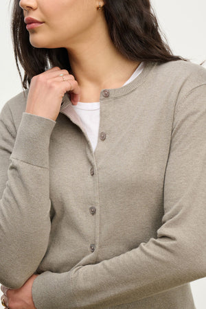 A woman with dark hair down touches her chin while wearing the VIOLETTE CARDIGAN by Velvet by Graham & Spencer in light beige cotton cashmere over a white shirt.