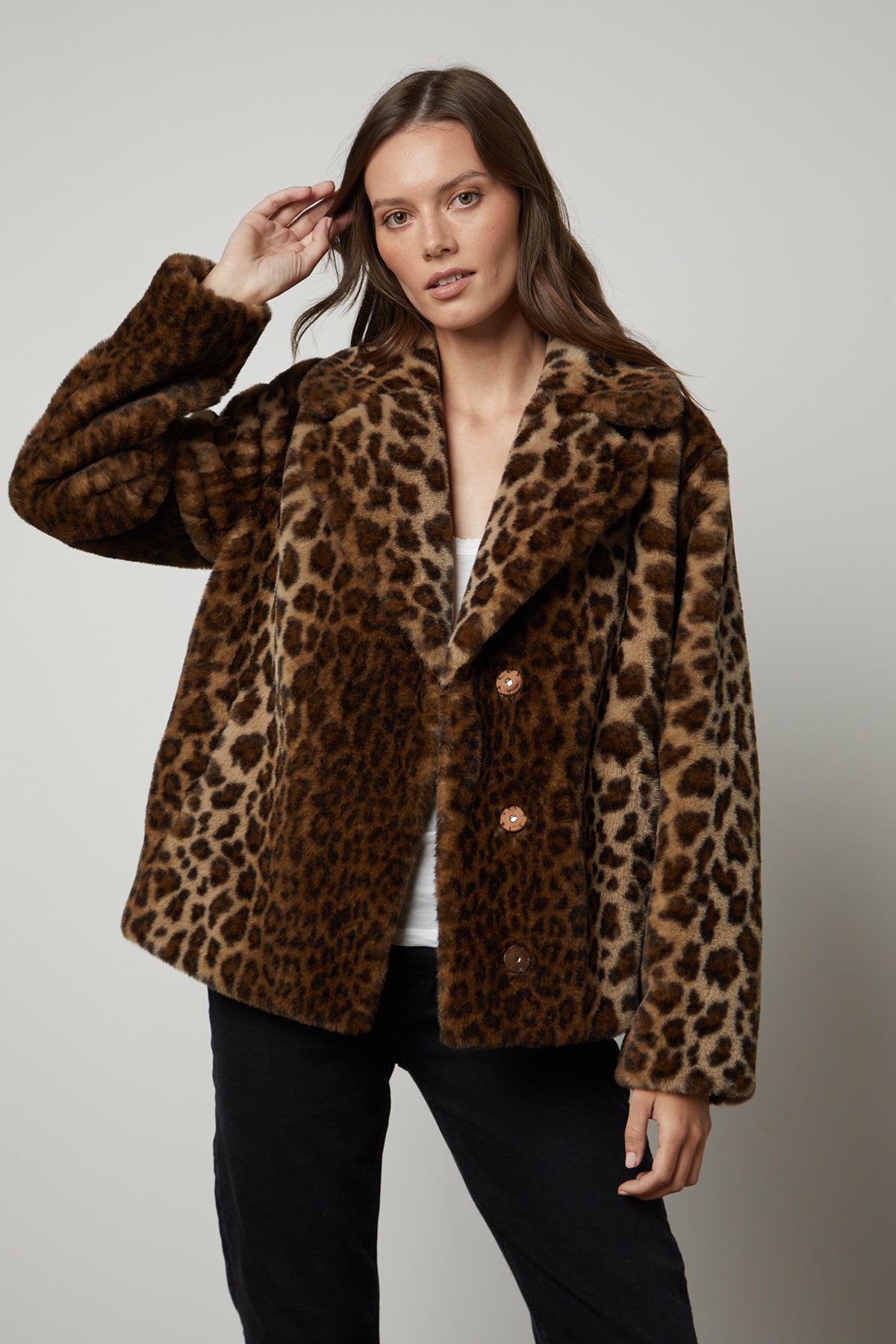 A woman wears a Velvet by Graham & Spencer AMANI LEOPARD LUX FAUX FUR JACKET, perfect for a fall wardrobe, and poses against a plain background. She has one hand raised to her head.
