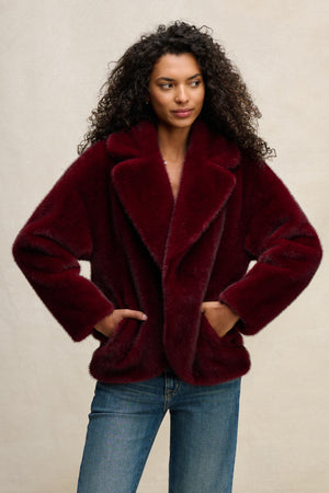 A woman with curly hair wears the Velvet by Graham & Spencer CAROLYN FAUX FUR JACKET in burgundy with a lapel collar and blue jeans, standing hands-in-pockets against a plain background.