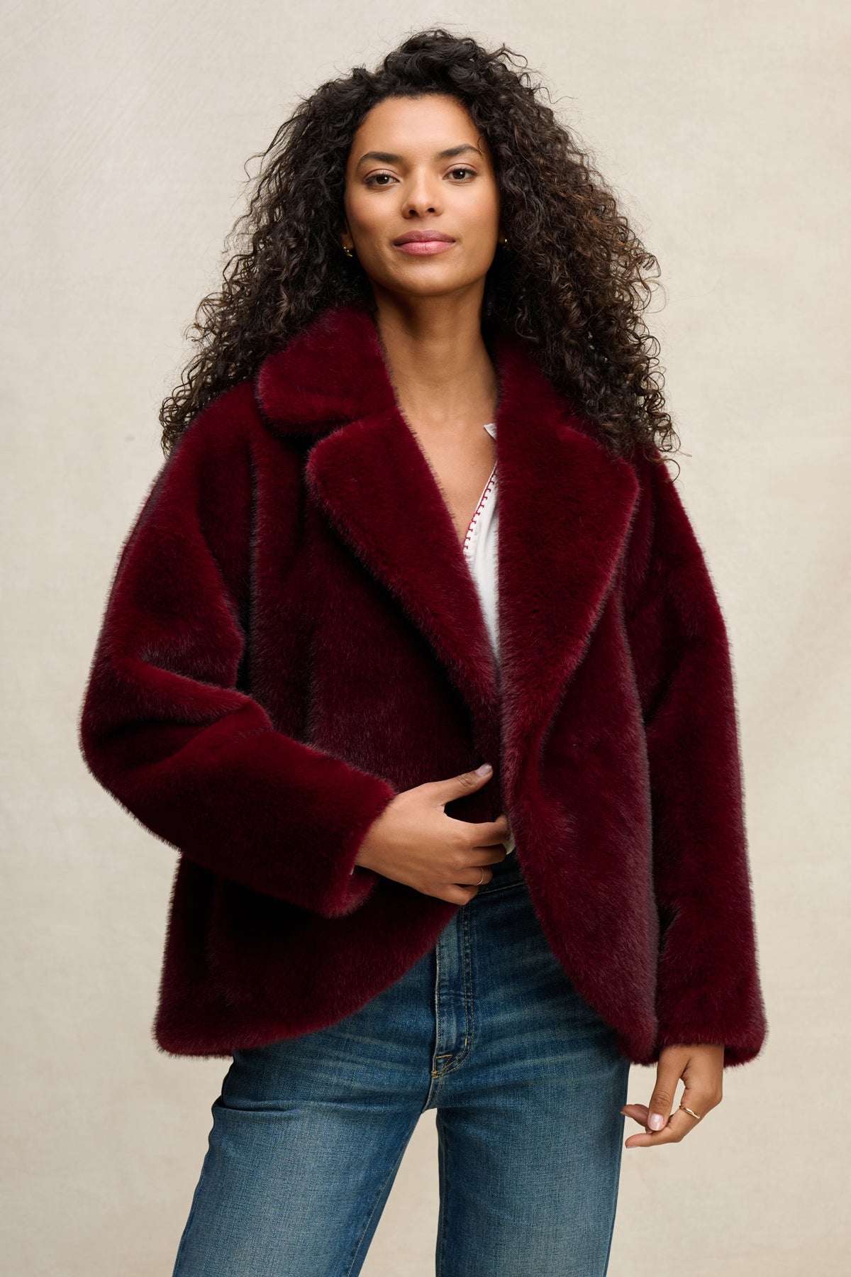 A person wears the CAROLYN FAUX FUR JACKET by Velvet by Graham & Spencer—a deep red faux fur jacket with a lapel collar—over a white top and blue jeans, standing against a plain light background.-39815134052545