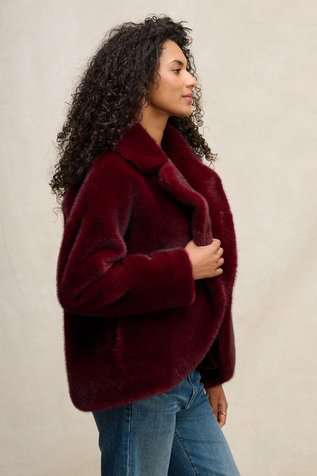 A woman in the CAROLYN FAUX FUR JACKET by Velvet by Graham & Spencer, featuring a lapel collar and burgundy faux fur, pairs it with blue jeans and stands side-on against a neutral background, touching the jacket's lapel.-39815134019777