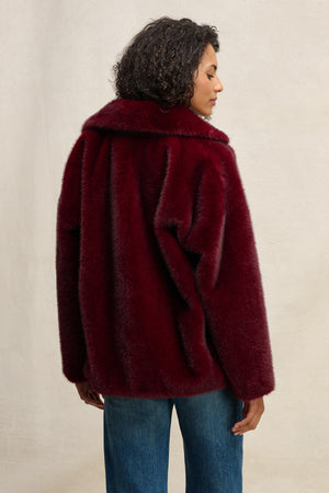 The CAROLYN FAUX FUR JACKET by Velvet by Graham & Spencer features a dark red hue, lapel collar, and plush texture—shown from the back with blue jeans against a light background—ideal for stylish outerwear lovers.