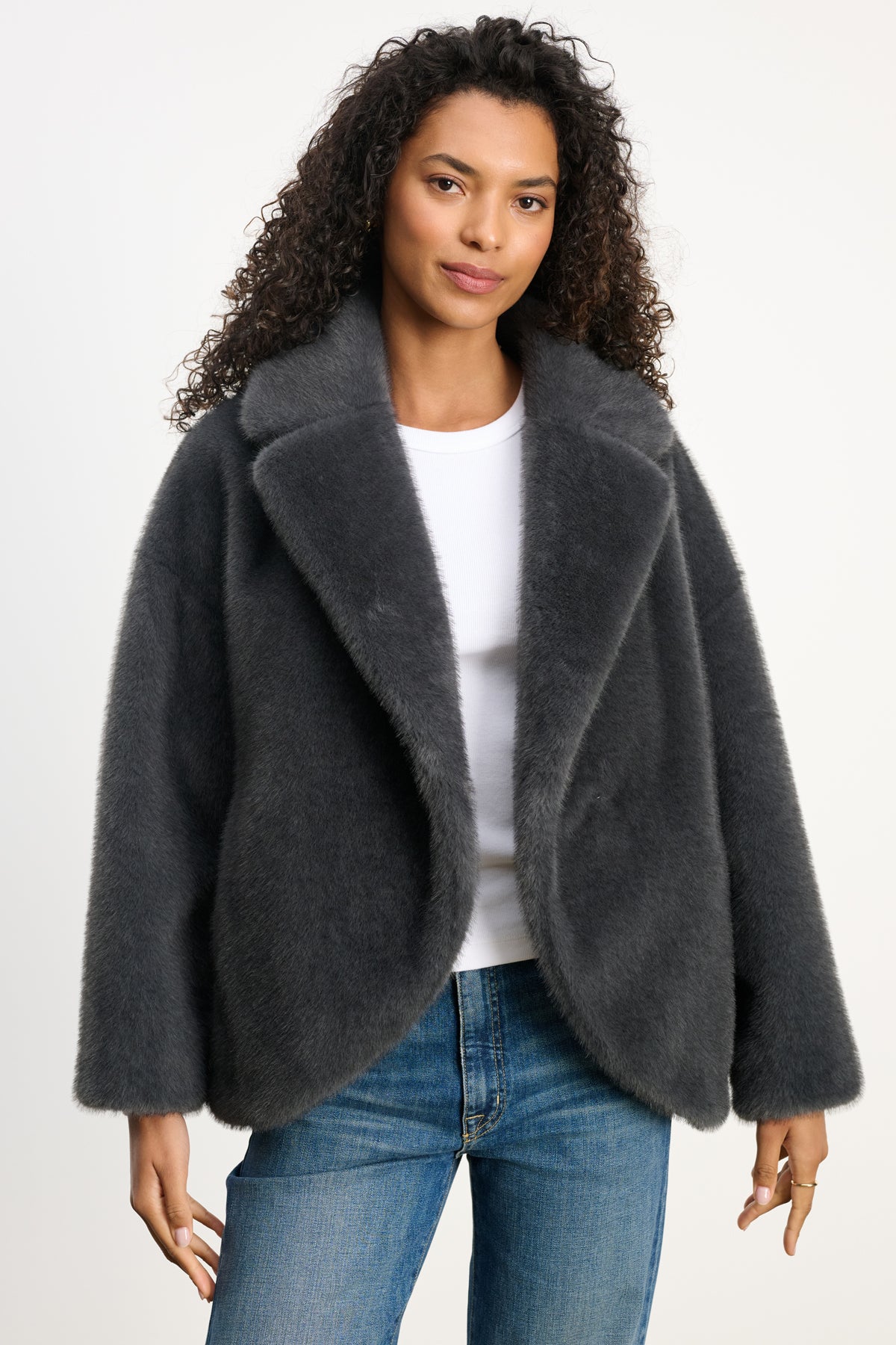 A woman wears the CAROLYN FAUX FUR JACKET by Velvet by Graham & Spencer, featuring a lapel collar, over a white shirt and blue jeans, standing against a plain light background.-39815133921473