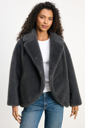 A woman wears the CAROLYN FAUX FUR JACKET by Velvet by Graham & Spencer, featuring a lapel collar, over a white shirt and blue jeans, standing against a plain light background.