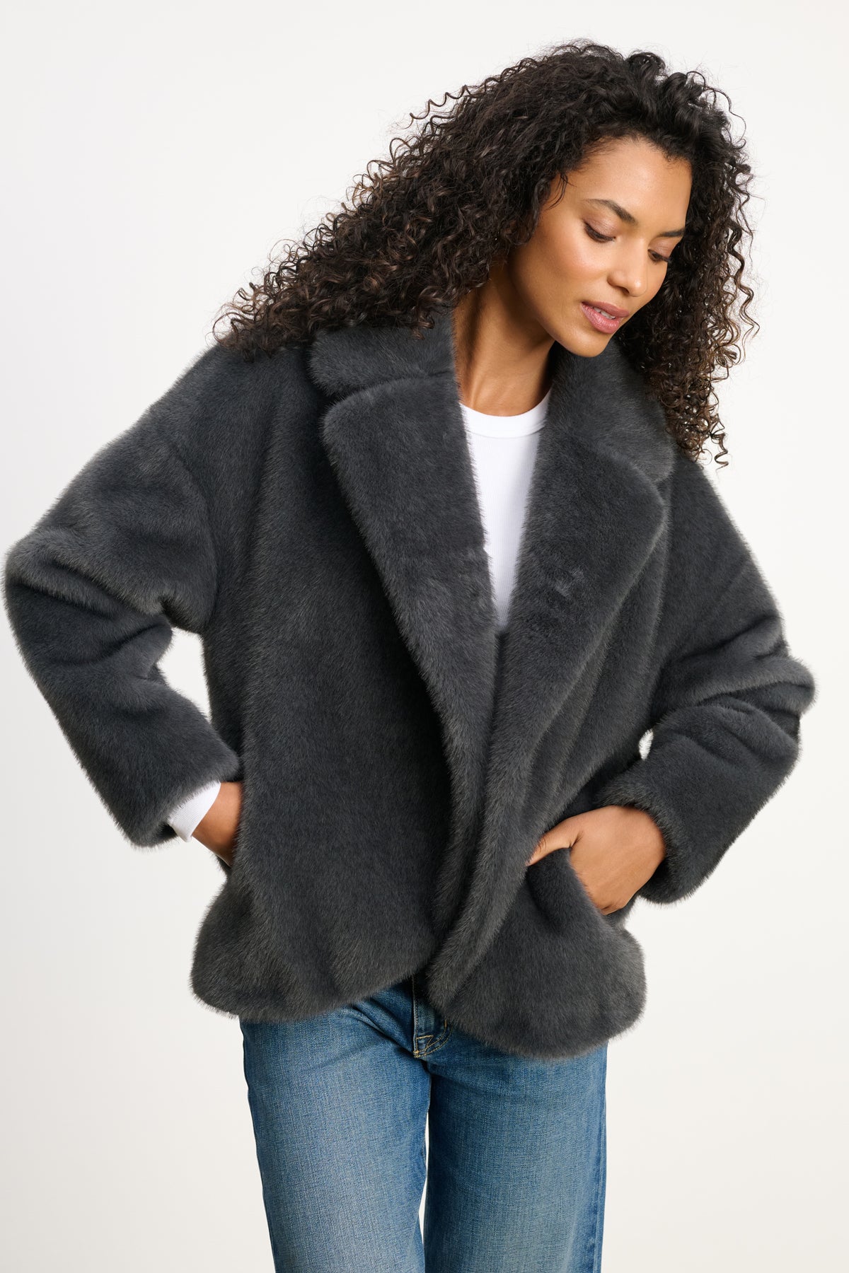 A woman wears the CAROLYN FAUX FUR JACKET by Velvet by Graham & Spencer, featuring a dark gray faux fur design with a lapel collar, over a white top and blue jeans, standing hands in pockets against a plain background.