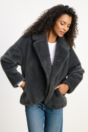 A woman wears the CAROLYN FAUX FUR JACKET by Velvet by Graham & Spencer, featuring a dark gray faux fur design with a lapel collar, over a white top and blue jeans, standing hands in pockets against a plain background.