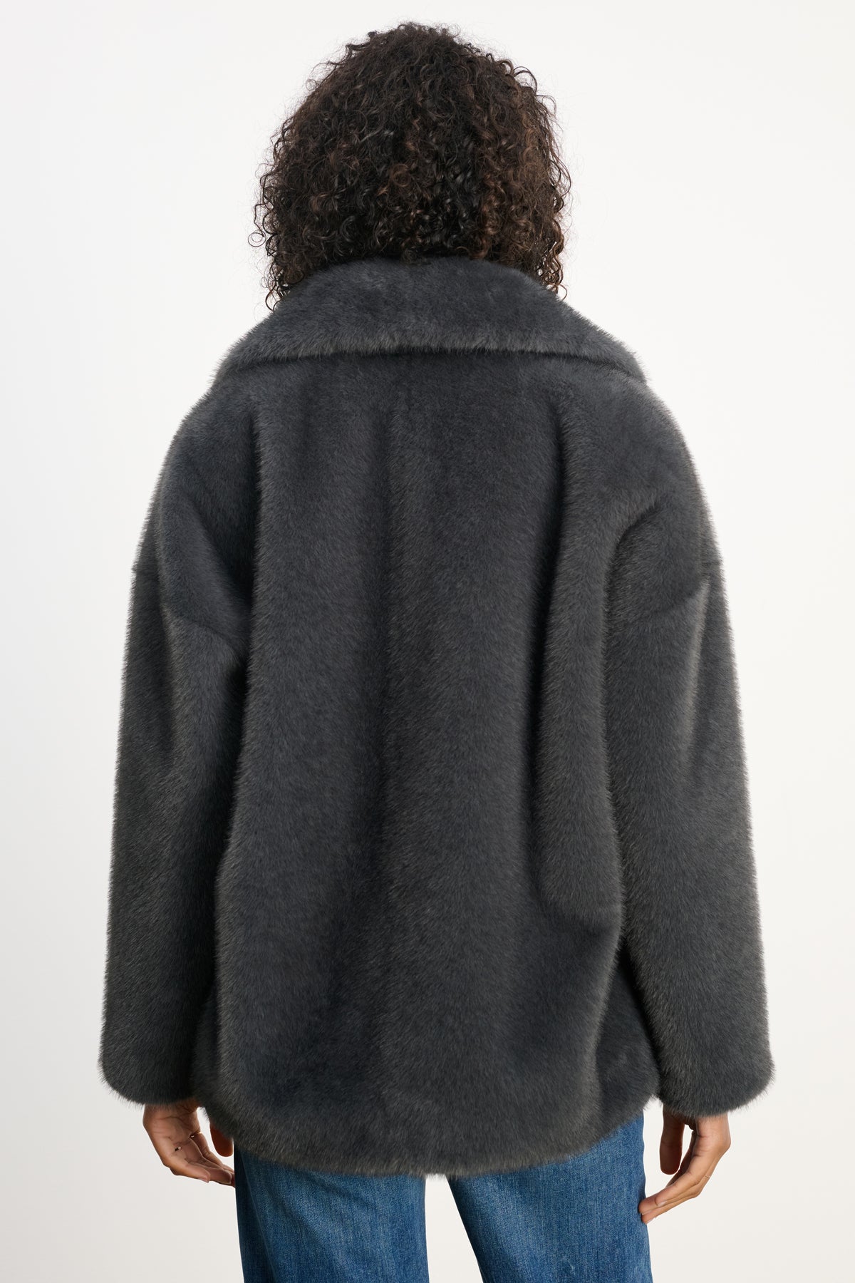 A person with curly hair wears the CAROLYN FAUX FUR JACKET by Velvet by Graham & Spencer—dark gray with a lapel collar—paired with blue jeans, shown from the back against a plain background.-39815133790401