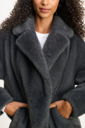 Model wears the CAROLYN FAUX FUR JACKET by Velvet by Graham & Spencer, a dark gray faux fur coat with a lapel collar, shown from shoulders to mid-thigh with hands in pockets.