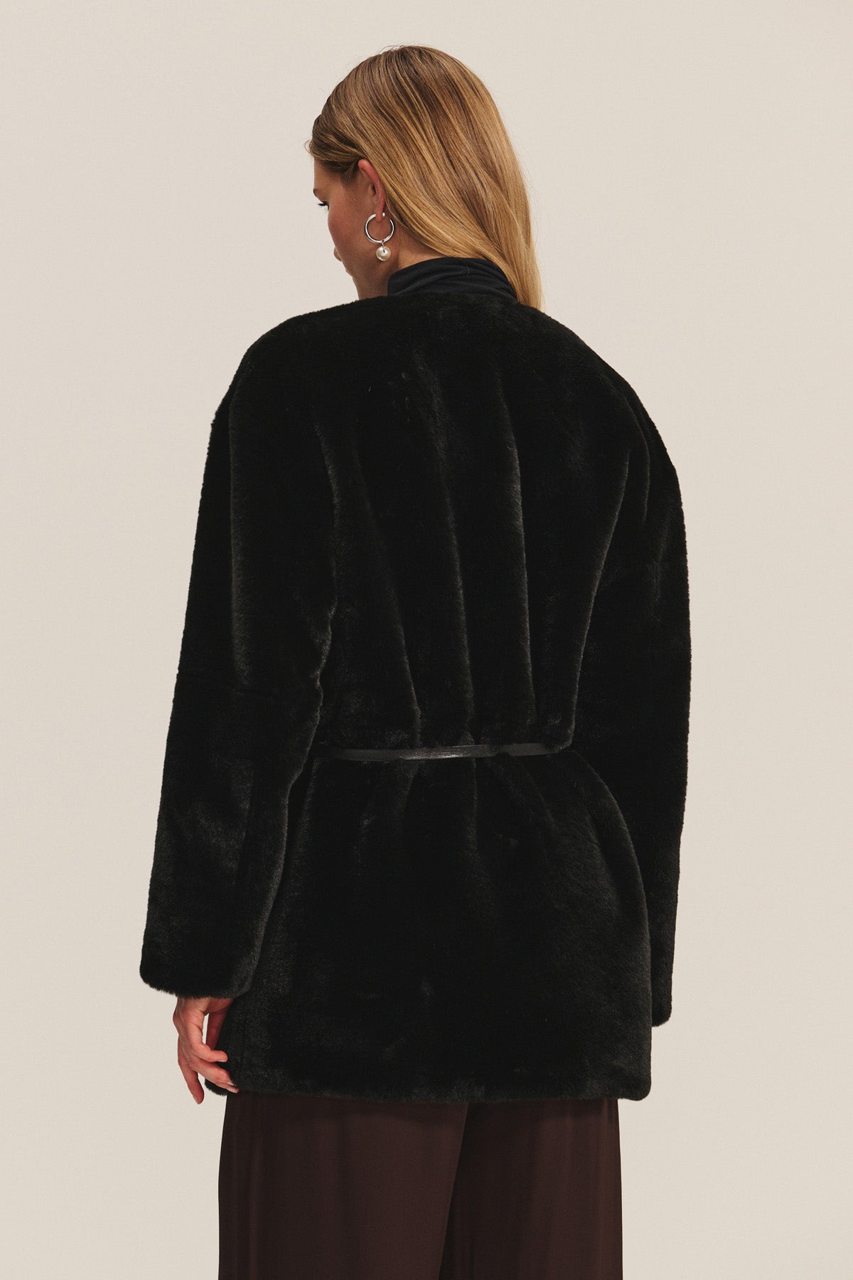 A woman with blonde hair is seen from behind wearing the MARMONT LUXE FAUX FUR JACKET by Velvet by Graham & Spencer, featuring snap button closures and dark pants against a light background.-40248194629825
