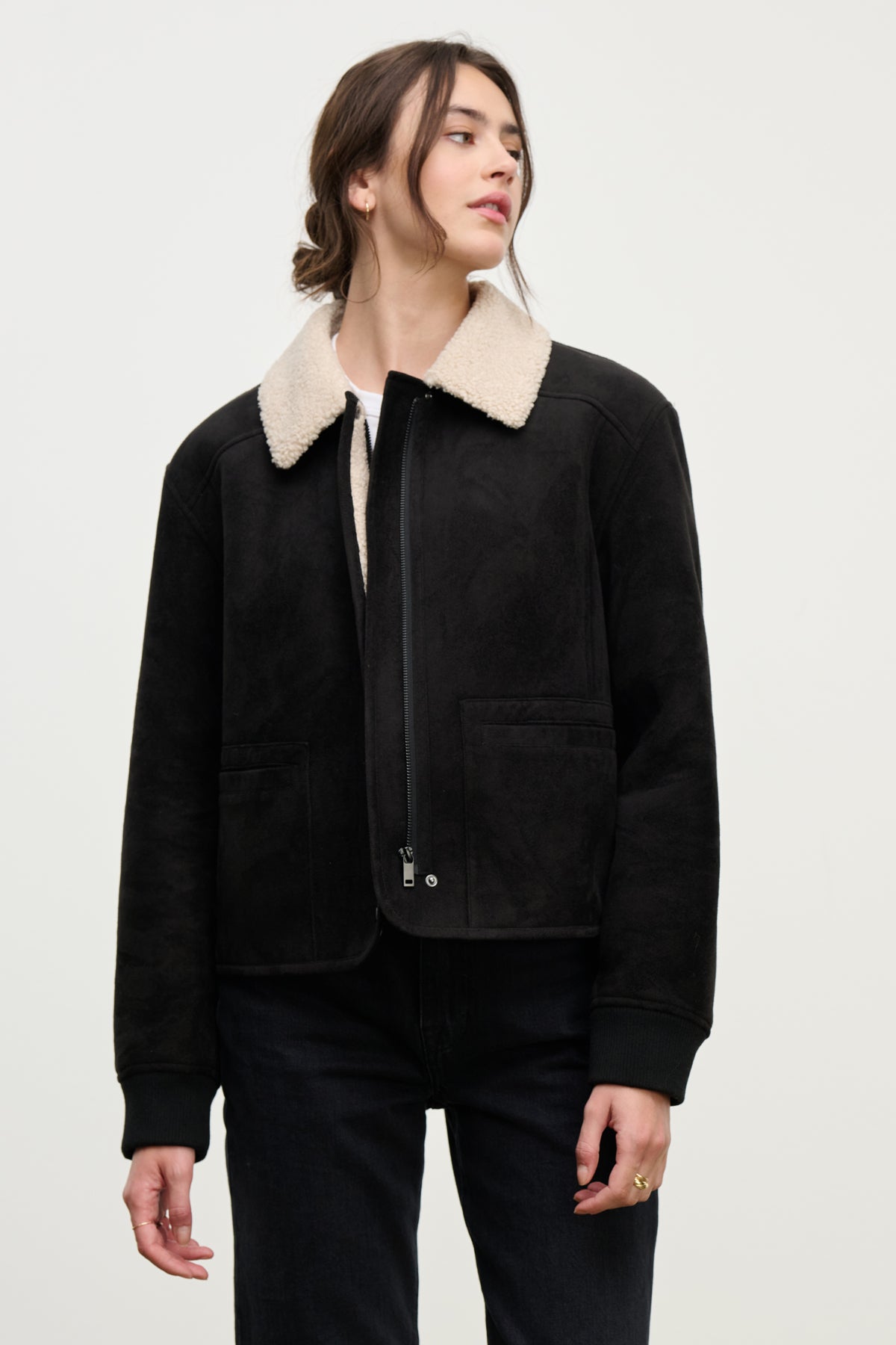 A woman wearing the JUNO SHERPA JACKET by Velvet by Graham & Spencer, featuring a black faux sherpa design and full zip front, pairs it with black pants and stands against a plain light background, looking to the side.-39838352900289