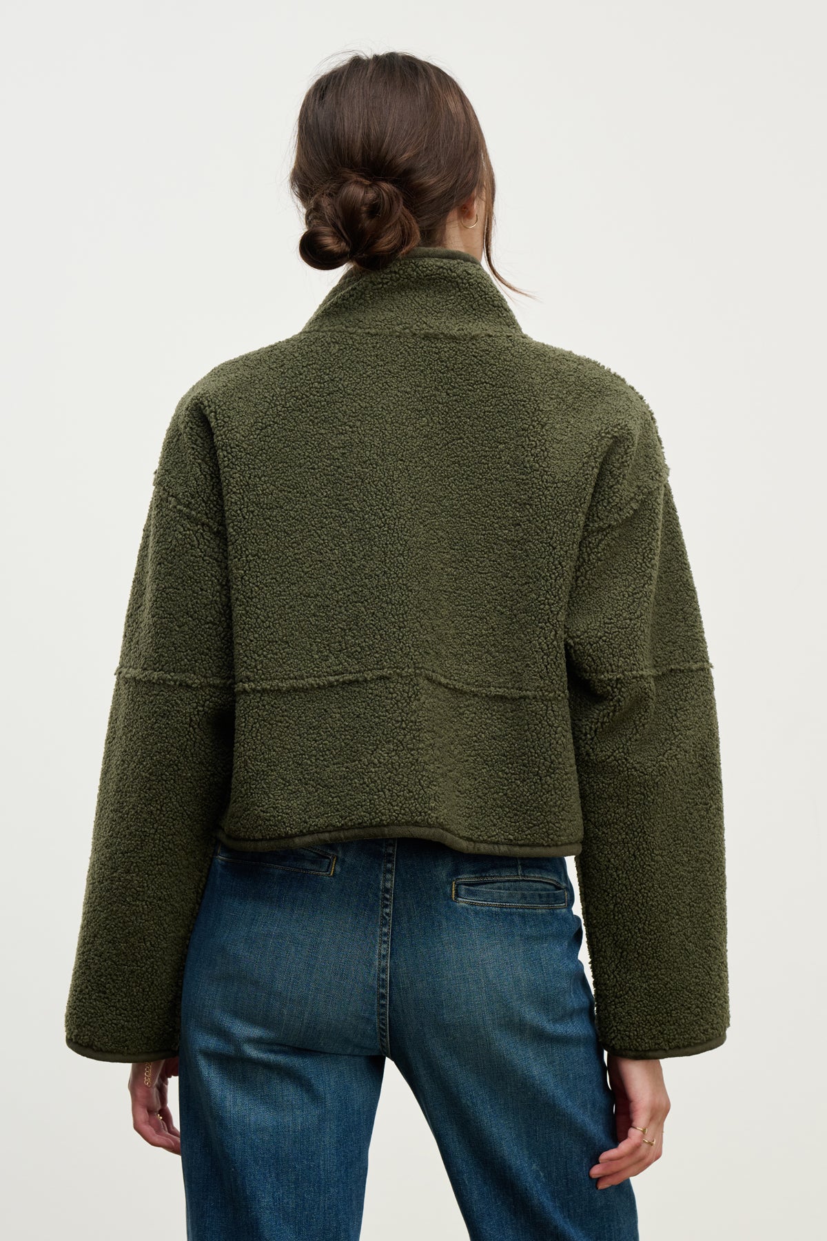 A person with brown hair in a low bun is seen from behind wearing Velvet by Graham & Spencer's KELLY LUXE SHERPA REVERSIBLE JACKET in olive green and blue jeans, set against a plain background.
