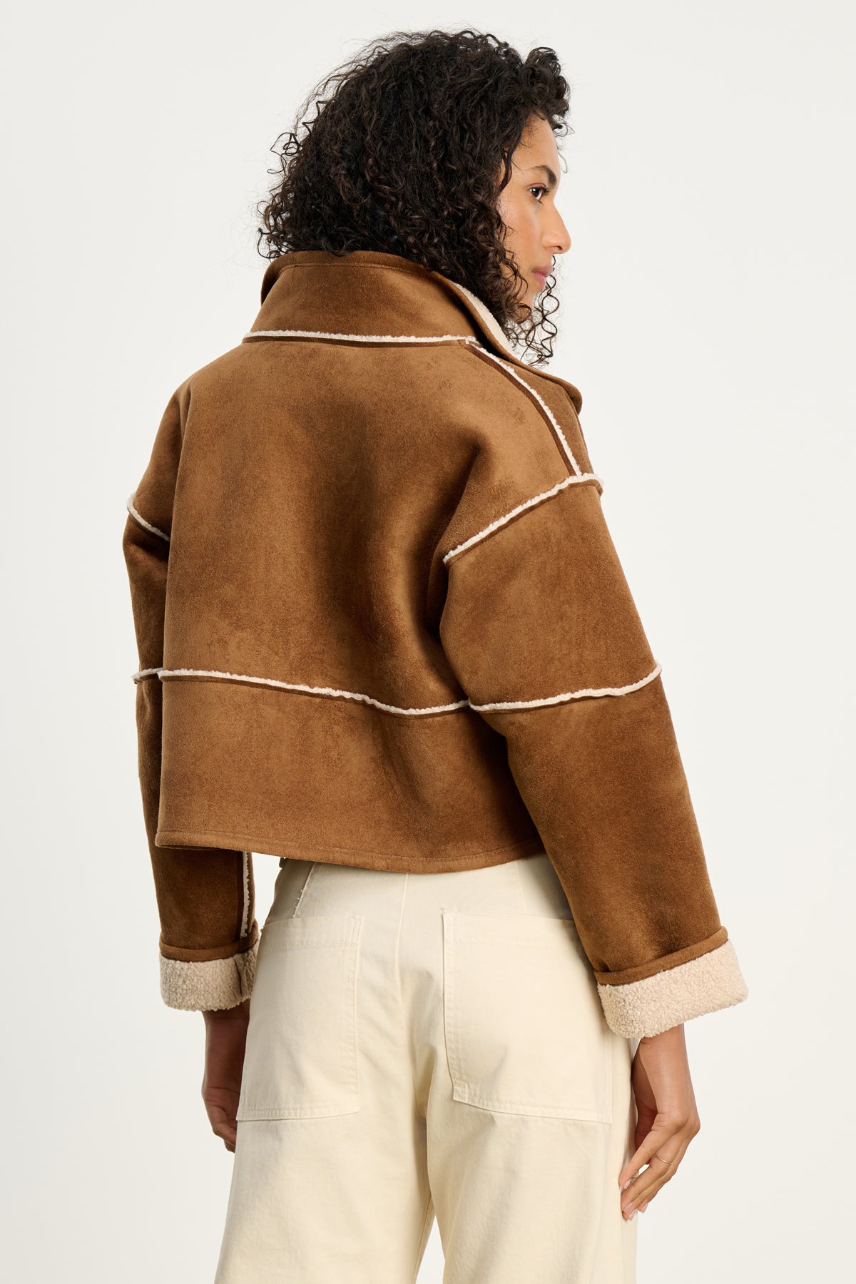 A woman with curly hair is shown from the back wearing Velvet by Graham & Spencer’s KELLY LUXE SHERPA REVERSIBLE JACKET, a brown cropped faux sherpa style with white trim, paired with cream-colored pants against a plain backdrop.