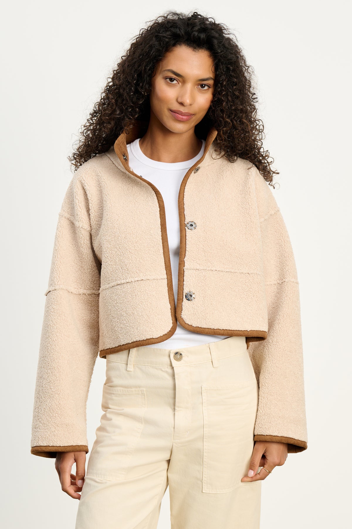 A woman stands against a plain background wearing the KELLY LUXE SHERPA REVERSIBLE JACKET by Velvet by Graham & Spencer, styled in beige with brown trim over a white tee and high-waisted cream pants.-39838356111553