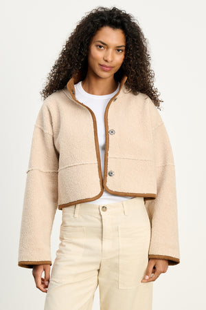 A woman stands against a plain background wearing the KELLY LUXE SHERPA REVERSIBLE JACKET by Velvet by Graham & Spencer, styled in beige with brown trim over a white tee and high-waisted cream pants.